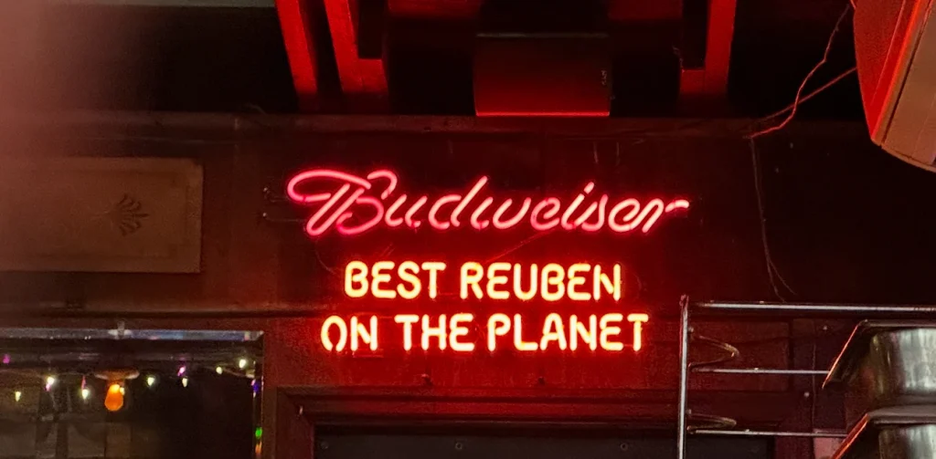 A red neon sign that says "Best Reuben on the planet." Above it sits a red neon Budweiser sign.